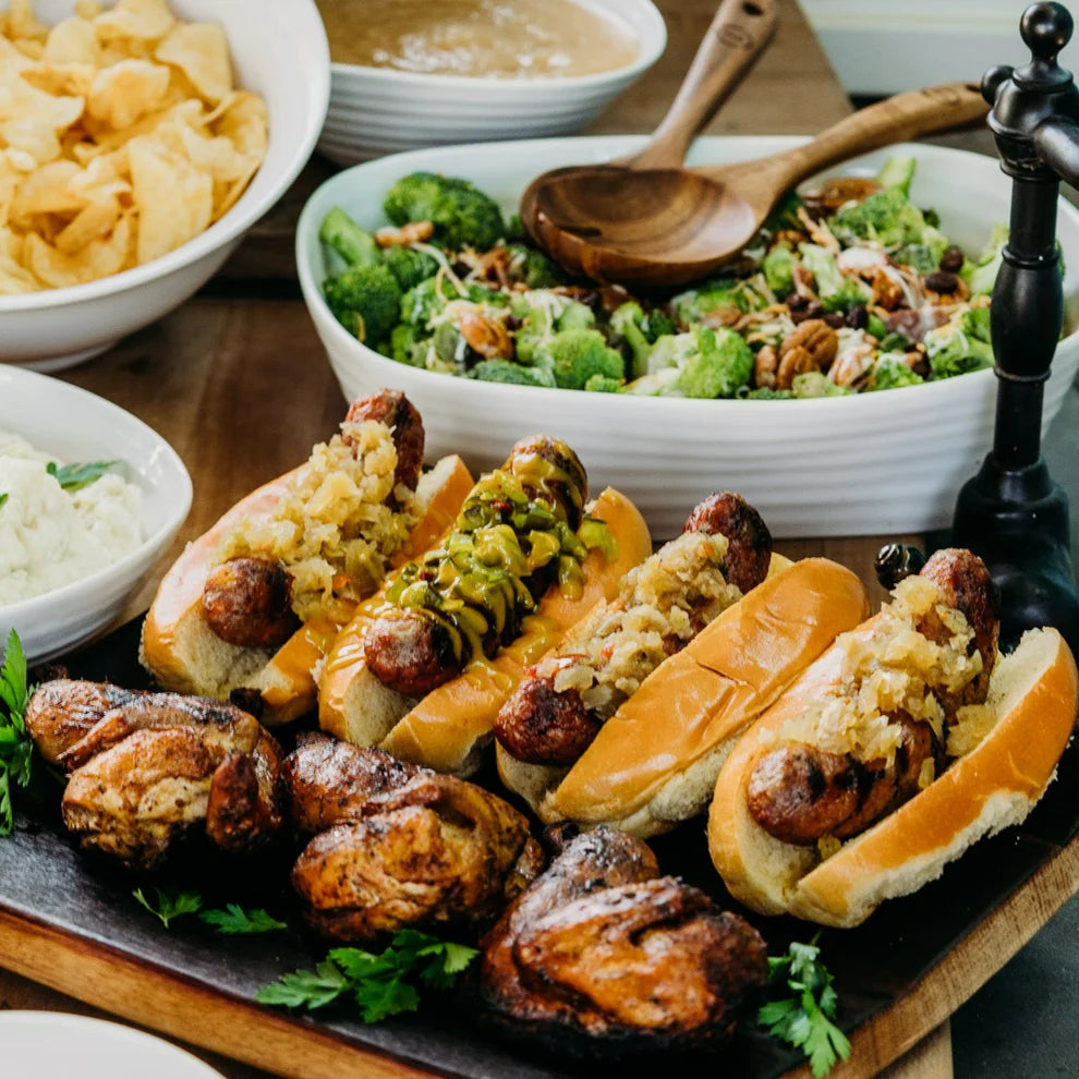 Ellis Bros Pecans Chow Chow on top of grilled brats on wooden board with chicken halves and broccoli salad